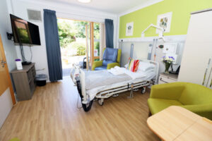 A bright hospital room with a single bed, colorful pillows, and a gray blanket. The room has light wood flooring, a TV, and a large window showing a garden. Green walls and chairs add a calming atmosphere.
