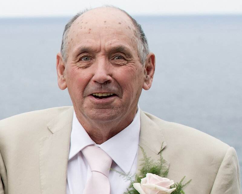 Man in a beige suit and pink tie with a boutonniere, standing outdoors near a body of water.