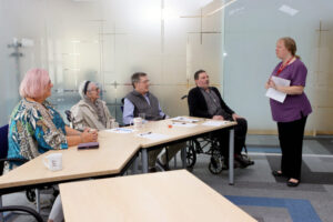 Group of seniors in wheelchairs attending a presentation by a standing instructor in purple uniform in a modern conference room.