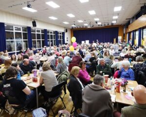 Large community event in a hall with many people seated at tables, colorful balloons, and a stage with a blue curtain in the background.