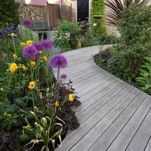 An view of a garden with multi coloured flowers and a wooden decking pathway leading into the distance