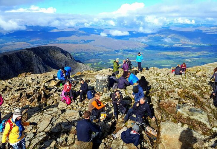 A diverse group of individuals atop a mountain, smiling and taking in the stunning scenery below