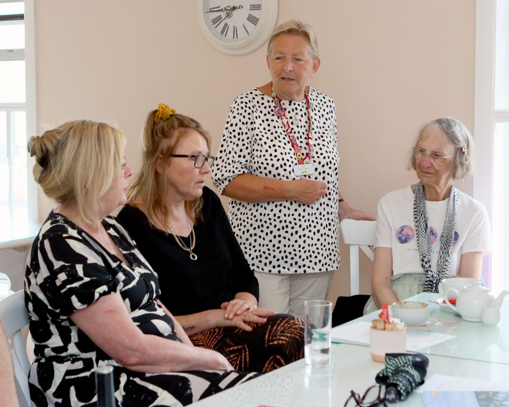 Adults sitting and standing around a table in a bright room, taking part in a bereavement help‑point conversation.