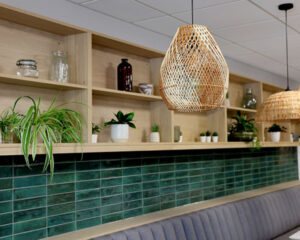 Modern interior with wooden shelves displaying plants and vases, green tiled backsplash, gray seating, and woven pendant lights hanging from the ceiling.