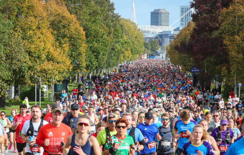 A large group of runners participates in the Cardiff half marathon on a tree-lined city street, with buildings visible in the background under a clear sky.