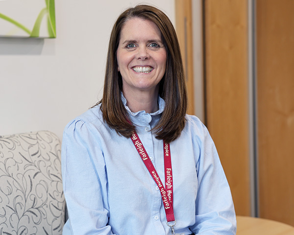 Person with long brown hair wearing a light blue blouse and a red lanyard, sitting on a patterned sofa in an indoor setting.