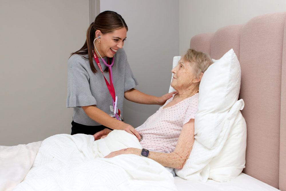 Clinical Nurse Specialist listening to an elderly patient's chest with a stethoscope in a home care setting.