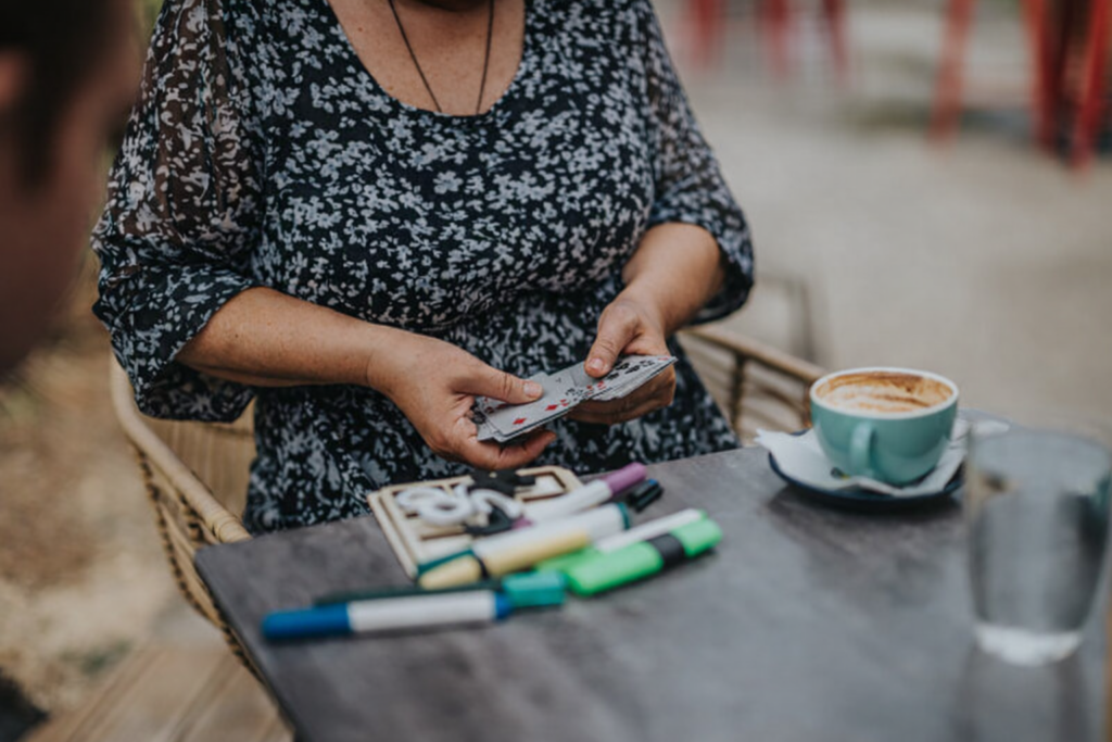 A person sits at a table holding playing cards during a patient and carer social drop‑in group. Coloured markers, a notebook, a coffee cup, and a glass of water are on the table, creating a relaxed, friendly atmosphere.