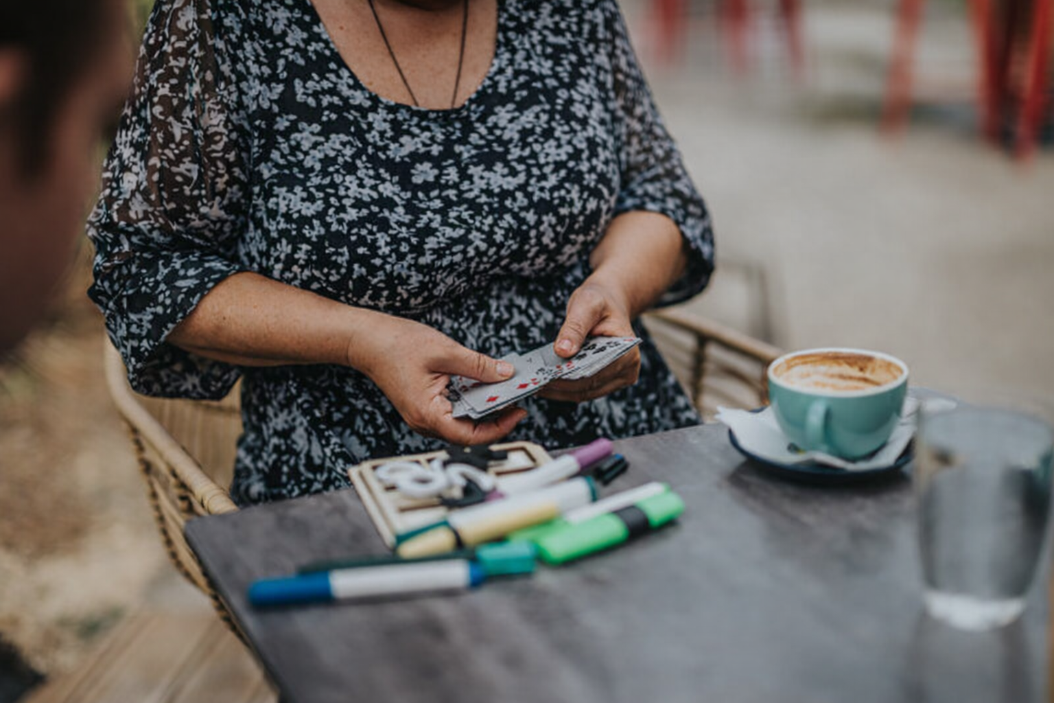 A person sits at a table holding playing cards during a patient and carer social drop‑in group. Coloured markers, a notebook, a coffee cup, and a glass of water are on the table, creating a relaxed, friendly atmosphere.
