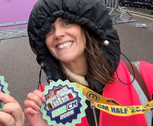 A brunette woman wearing a black coat with the hood up, holding up a medal with a big smile on her face.