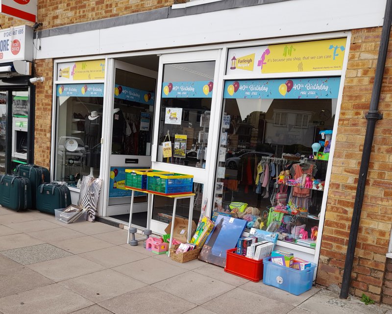 A charity shop front displaying colorful signage celebrating its 40th birthday, with various items for sale visible outside. Suitcases and toys are arranged on the pavement, inviting shoppers to explore.