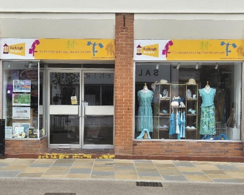 Facade of the Farleigh Hospice shop showcasing a variety of colorful women's clothing in the display windows. The shop's yellow sign includes a fundraising message and a logo.