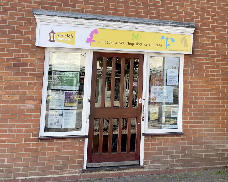 Front view of a brick building with a wooden door and windows displaying posters, featuring a yellow Farleigh Hospice shop sign above.