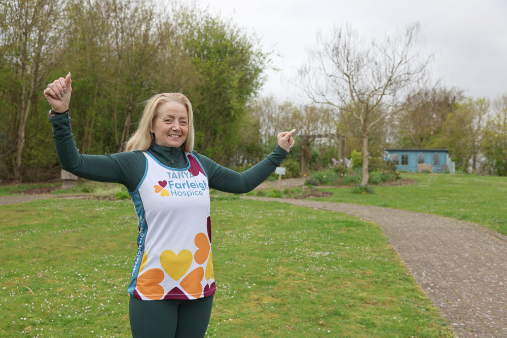 A woman stands in the garden of the hospice with arms aloft wearing a Farleigh branded running top