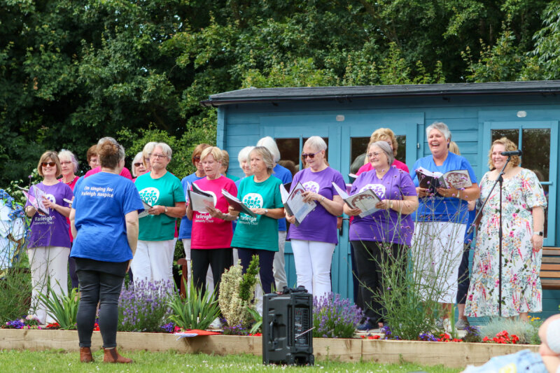 A group of people sing outdoors in front of a small blue wooden building as part of the Farleigh Hospice choir. They wear brightly coloured shirts with “We’re singing for Farleigh Hospice” printed on them, holding songbooks while a conductor leads the group. Flowers and trees surround the area, creating a cheerful community atmosphere.