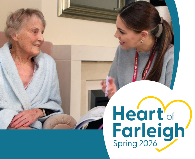 Smiling elderly woman in a light blue robe sits in a cosy living room, engaging warmly with a younger female caregiver wearing a gray top and ID lanyard, who is holding a notebook and gesturing as they chat; soft neutral décor with a fireplace and mirror in the background creates a calm, supportive care environment. Overlay text reads “Heart of Farleigh Spring 2026” with a stylized yellow heart graphic, suggesting a healthcare or hospice care campaign focused on compassion and connection.