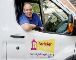 A smiling person in a blue shirt leans out of a white van window. The van has a Farleigh Hospice logo, conveying a friendly, charitable tone.