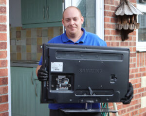 A person in a blue shirt and black gloves holds the back of a Samsung TV, standing in a kitchen with a tiled backsplash, conveying a feeling of helpfulness.