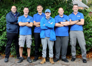 Six men in matching blue shirts stand smiling with arms crossed in front of lush green foliage