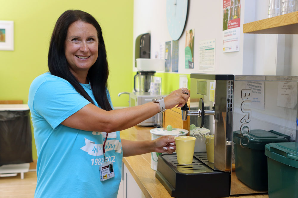 A smiling female wearing a Farleigh branded t-shirt stands at a coffee making machine