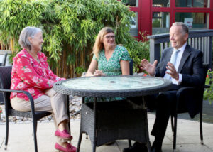 Three individuals engage in conversation around a circular table in a garden setting. Lush green plants provide a natural backdrop as they share smiles and gestures.