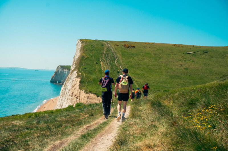 A scenic mountain view on a clifftop path with green grass and a bright blue sky, with a far away shot of people walking on a trail with hats and backpacks on.