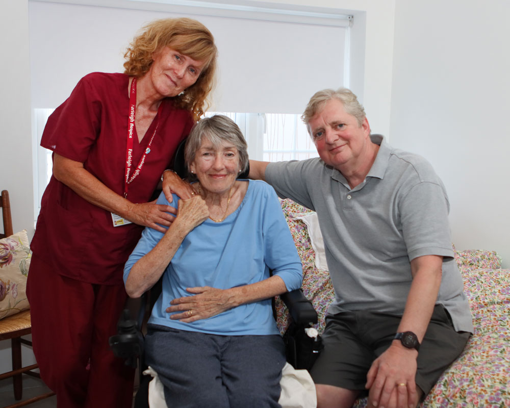 A person living with motor neurone disease sits in a wheelchair, supported by a nurse and her husband at home.