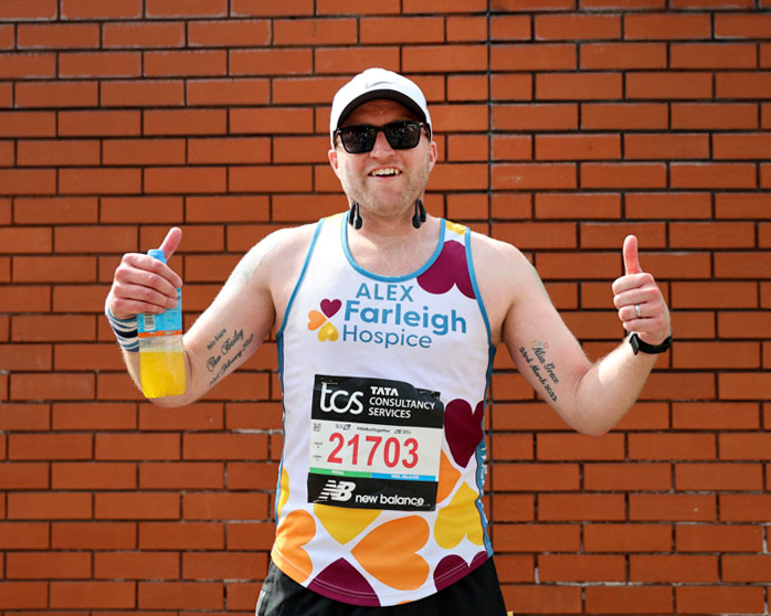 A male Marathon runner wearing a Farleigh branded running vest smiles at the camera with his thumbs up