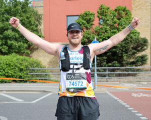 A male marathon runner wearing Farleigh Hospice branded top smiles as he holds his arms up on a London street.