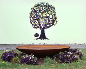 A flat metal tree tree sculpture is shown fixed to a wall with a shallow dish water feature in front surrounded by flowers