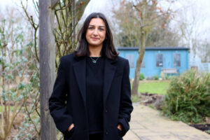 A woman stands outdoors in a black coat, smiling warmly in front of a blue building surrounded by greenery. The scene conveys a relaxed, natural environment.