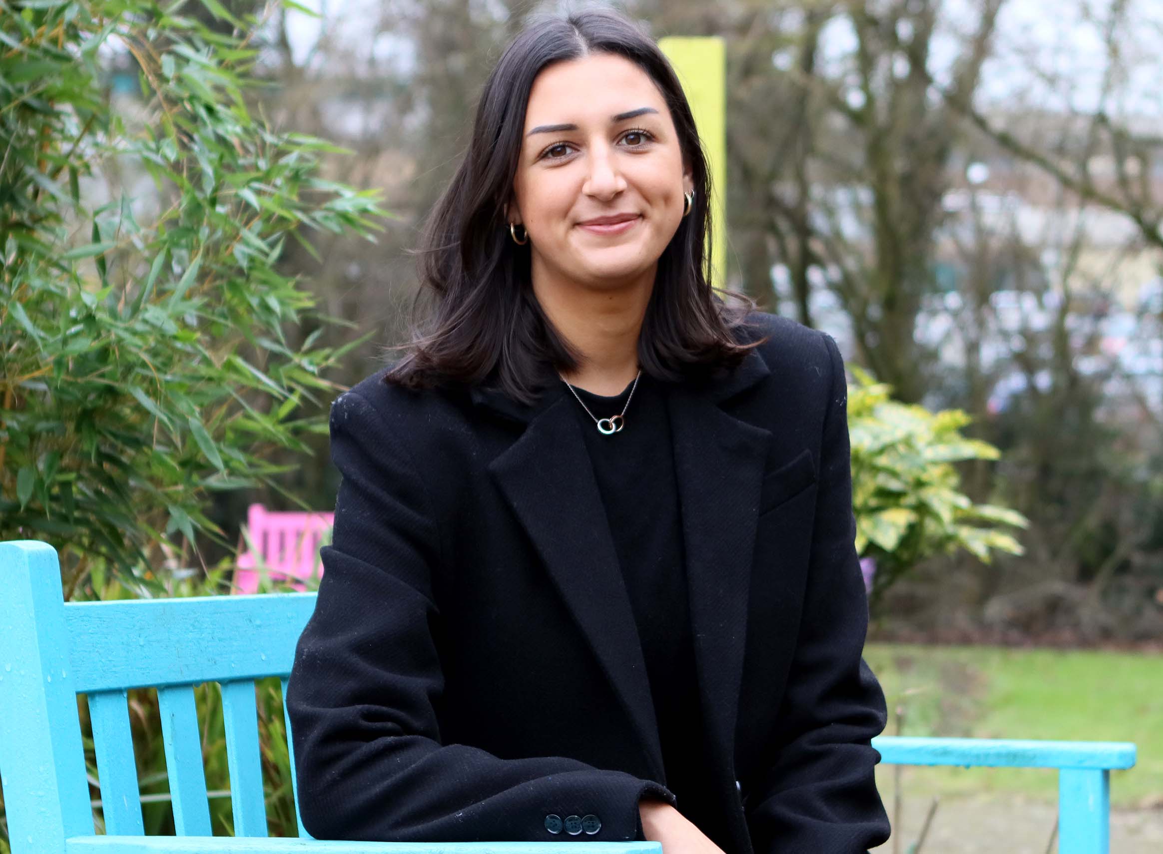 A person with shoulder-length dark hair, wearing a black coat, sits on a blue bench outdoors with greenery and trees in the background.