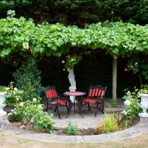 A bistro style table and chairs under a pergola with flowers in a round flower bed