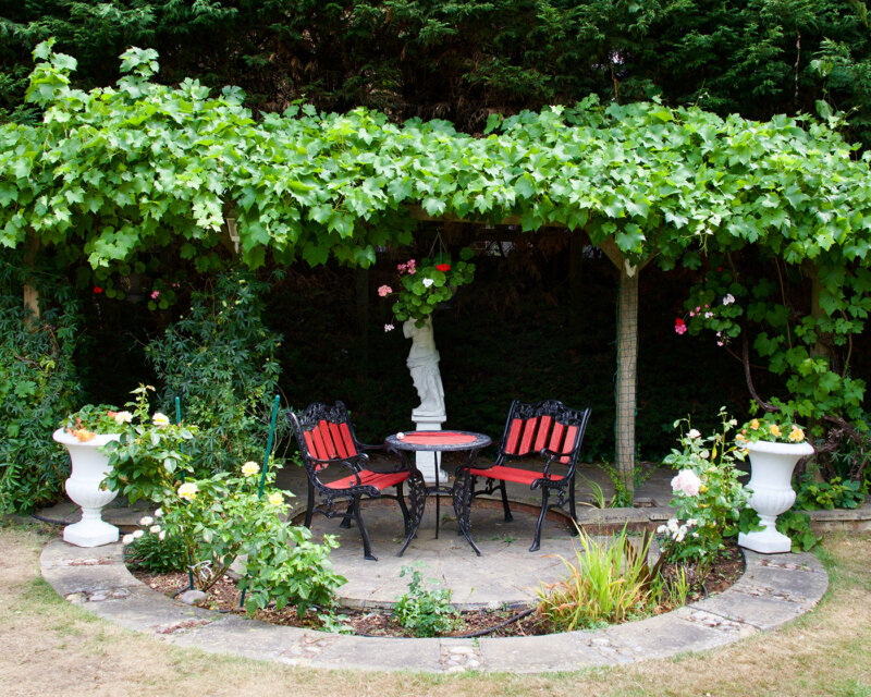 A bistro style table and chairs under a pergola with flowers in a round flower bed