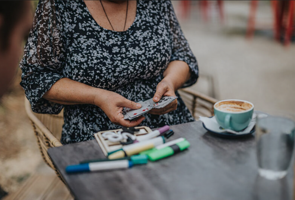 A person sits at a table holding playing cards during a patient and carer social drop‑in group. Coloured markers, a notebook, a coffee cup, and a glass of water are on the table, creating a relaxed, friendly atmosphere.