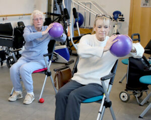Two older adults sit in a rehabilitation room holding purple exercise balls with their arms extended forward, performing a seated strength exercise. Wheelchairs and therapy equipment are visible in the background.