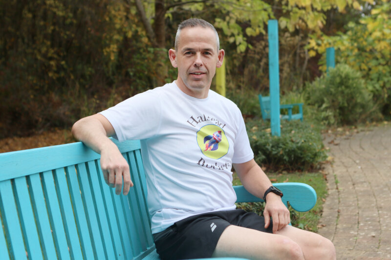 Man in a white T-shirt with a rocket logo sits on a bright turquoise park bench surrounded by autumn foliage.