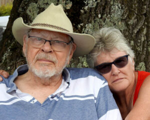 Elderly couple sitting outdoors by a tree, the man wearing a cowboy hat and a striped polo shirt, the woman in a red sleeveless top.