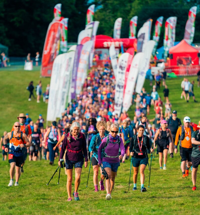 A big group of people with walking gear and backpacks walking along a grassy field with branded flags surrounding them.