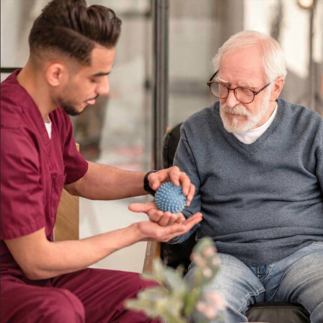 A Farleigh Hospice physiotherapist wearing burgundy scrubs supports an older patient during a therapy session, demonstrating the use of a blue spiky massage ball to aid mobility and physical wellbeing in a calm, well-lit treatment room.
