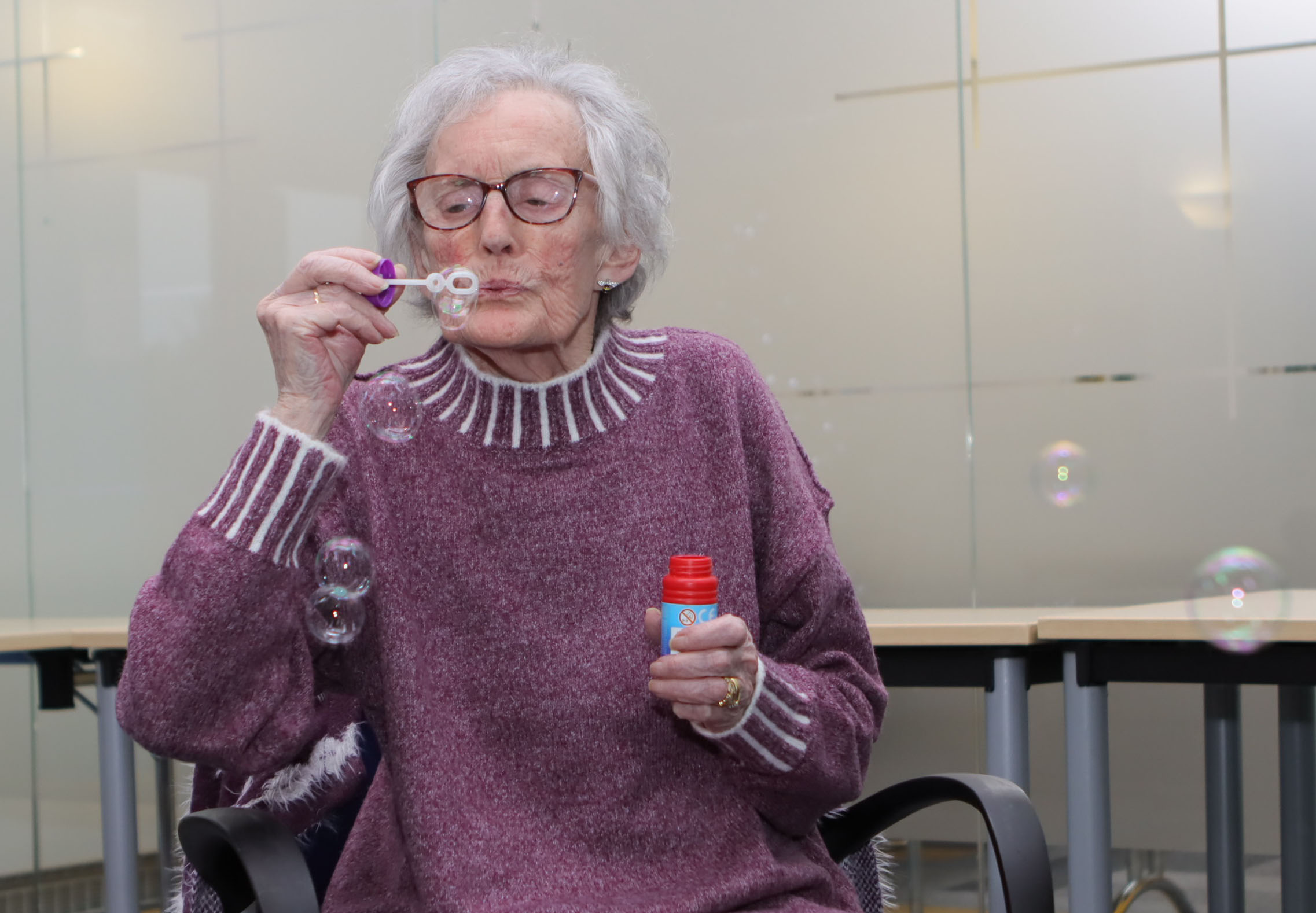 Elderly lady wearing a purple sweater blowing soap bubbles indoors, seated near a round table with a glass-paneled wall background.