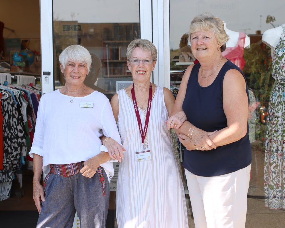 Three female retail volunteers stand outside a Farleigh shop with arms linked smiling at the camera