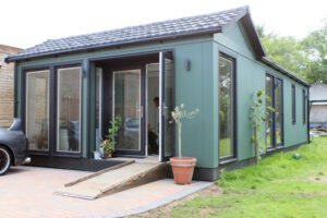 A modern green house with large glass windows and a sloped roof, featuring a front porch with steps leading up to the entrance. Decorative plants are placed near the door, and there is a small garden area in front of the house.