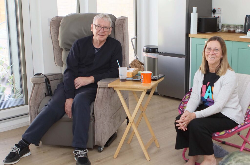 Two people sitting in a living space. One person, an older man in glasses, is sitting in a recliner chair with a drink and some snacks on a small table beside him. The other person, a woman with long hair wearing a colorful shirt, is seated on a folding chair next to the table.
