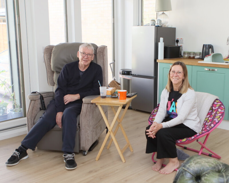 A man living with motor neurone disease sits at home with his wife beside him. They are seated comfortably in their living room, talking quietly together with drinks on the table between them. The scene shows a calm, moment that reflects how they manage life at home with support from Farleigh Hospice.