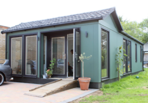 A single‑storey accessible home with a green exterior, large windows and a wooden ramp leading to the entrance. Potted plants sit by the doorway and a driveway runs alongside the house, reflecting the adapted home where Richard and Moira live.