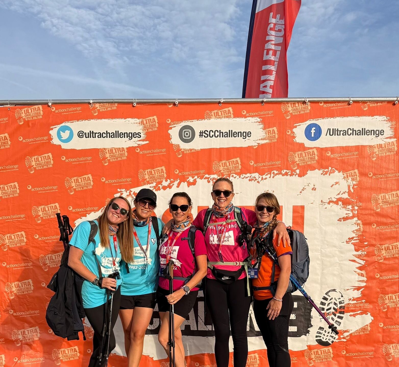 A group of five South Coast Ultra Challenge participants pose with trekking poles in front of the event backdrop while fundraising for Farleigh Hospice.