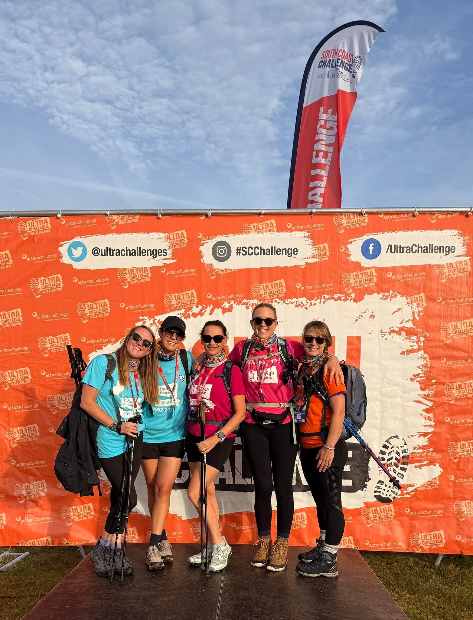 A group of five South Coast Ultra Challenge participants pose with trekking poles in front of the event backdrop while fundraising for Farleigh Hospice.