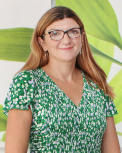A woman with long brown hair, wearing glasses and a green floral dress, stands in front of a backdrop with green leaves. Her expression is friendly and welcoming.
