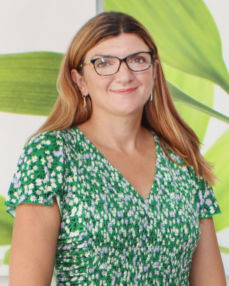 A woman with long brown hair, wearing glasses and a green floral dress, stands in front of a backdrop with green leaves. Her expression is friendly and welcoming.